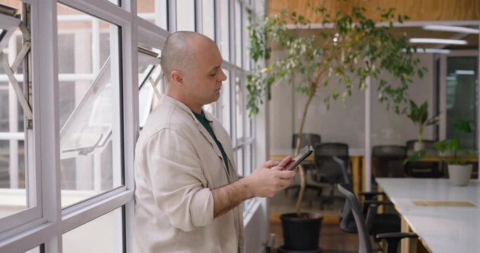 Adult man leaning by office panes glancing right, tapping tablet while checking work wearing band