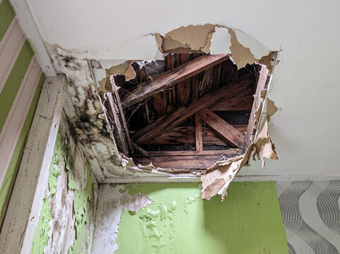 Damaged drywall ceiling hole in house interior exposing wooden roof structure frame needing repair and renovation