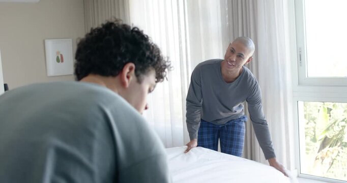 African American men pulling, smoothing white duvet in bedroom wearing grey PJs, by sun