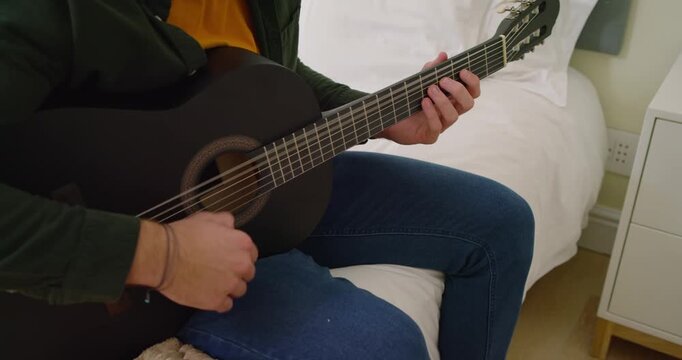 Close-up showing man in green shirt, jeans fretting and strumming black guitar on bed to relax