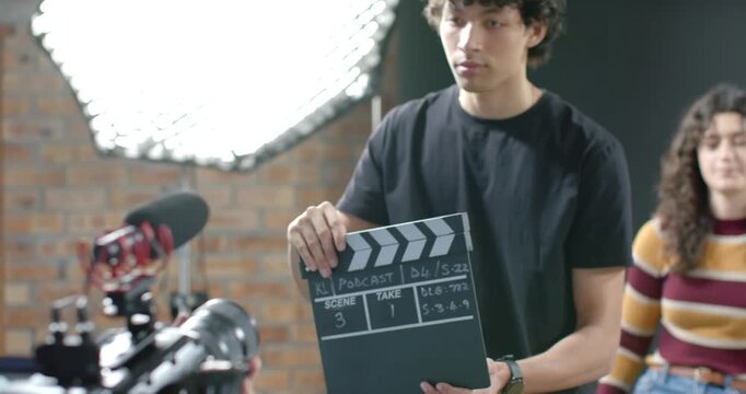 Adult male crew clapping clapperboard, marking KL Podcast set, female talent sitting facing camera