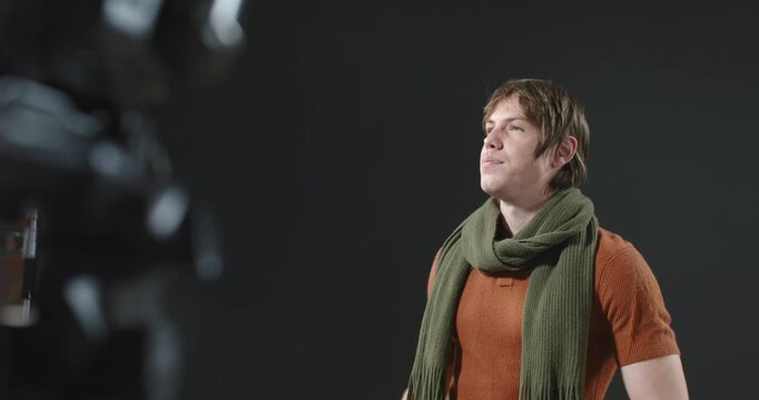 Male actor responding to off-camera cue, shifting to studio stool, tightening scarf