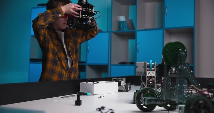 Young person assembles and lifts a small robot prototype while examining exposed mechanical components at a workshop table in a robotics laboratory