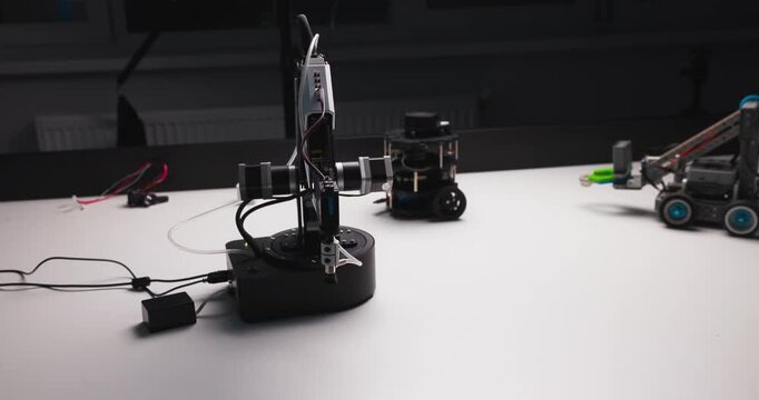 Robotic arm rotates and adjusts position beside small wheeled robots during a tabletop demonstration in a dark laboratory