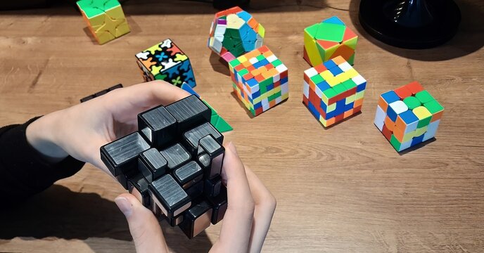A boy&rsquo;s hands holding a Mirror Blocks puzzle cube (QiYi model); in the background, other geometric and mechanical puzzles are visible, such as a Megaminx (dodecahedron-shaped) and various other speed 