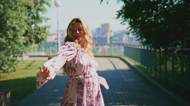 Sunlit park path with green trees lining both sides. Woman in floral dress walks barefoot with open arms, smiling. City skyline visible in soft focus beyond the fence