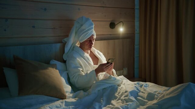 Cozy bedroom scene with soft lighting. Person wrapped in towel, relaxed, absorbed in phone. Warm wooden walls, gentle glow from bedside lamp. Comfortable bed with plush pillows, white duvet