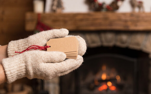 Hands in woolen mittens holding blank paper tags with red string for Christmas gifts near fireplace