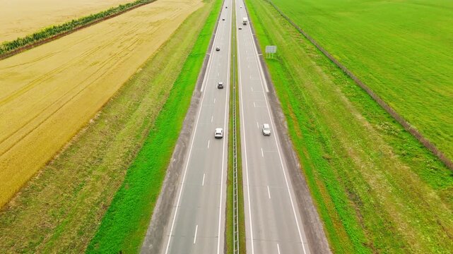 Aerial view of highway cutting through fields. Vehicles travel along the road. Green and golden fields flank the asphalt. Road stretches into the horizon. Perfect for travel or transport themes