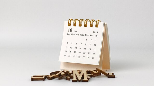 A small spiral-bound October 2026 desk calendar stands behind a scattered pile of wooden letters on a white surface.