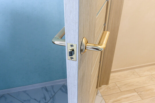 Light‑wood interior door with brushed gold lever handle, visible latch plate, clean panel lines, set against pale blue wall and tiled floor