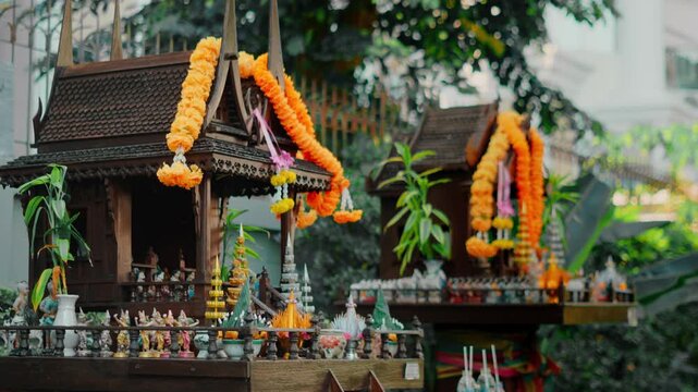 Ornate wooden spirit houses in Thailand adorned with bright flower garlands, religious offerings. Traditional miniature shrines reflect spiritual beliefs, cultural identity, centuries local customs.