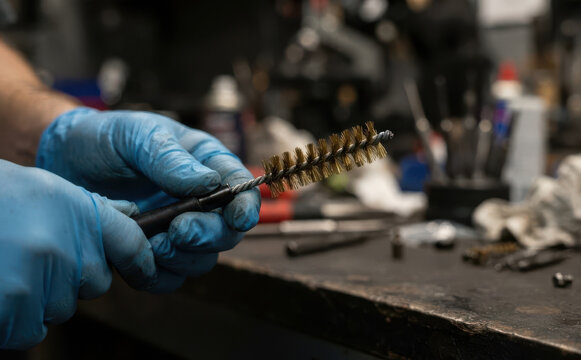 Gloved hands holding a brass cleaning brush for firearms maintenance in a workshop setting representing gun care and gunsmithing expertise