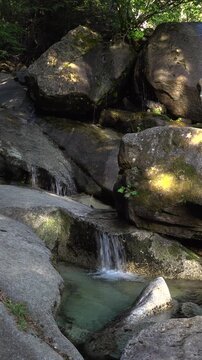 A vertical stationary shot of fresh water trickling and cascading over large granite boulders. Sunlight highlights the dark stone textures during a summer day at the bottom of Shannon Falls.