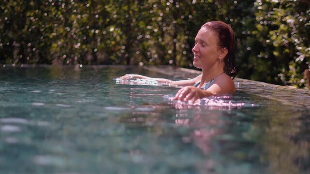 Relaxed woman enjoys peaceful moments in tropical swimming pool, leaning against pool edge with closed eyes. Warm sunlight, lush greenery, refreshing water create perfect summer vacation atmosphere.