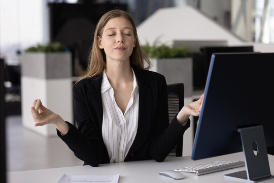 Mindful young female business professional in formal suit meditating at office desk practicing yoga breathing exercises for workplace relaxation stress relief mental wellness productivity improvement