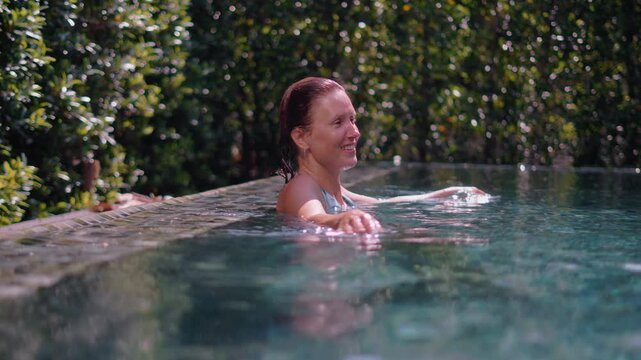 Young woman rests comfortably against swimming pool edge, enjoying warm sunshine, tranquil tropical surroundings. Gentle smile, closed eyes express happiness, relaxation and carefree summer leisure.