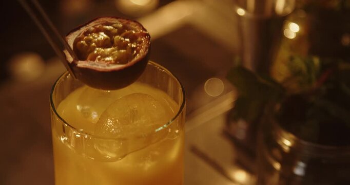 Close up of a bartender holding a halved passion fruit with metal tongs above an orange cocktail containing a large ice sphere. Warm golden lighting highlights the juicy pulp and elegant presentation.