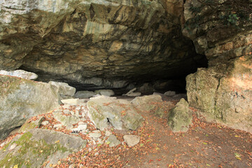 A spacious limestone cave entrance with low ceilings and scattered boulders on the forest floor. Autumn leaves enhance the sense of isolation and mystery. © Pro Photo Factory