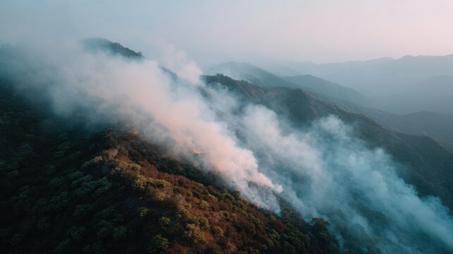 A drone surveys wildfire spreading across a mountain ridge, where smoke drifts with the wind above burnt trees and forest under atmospheric haze