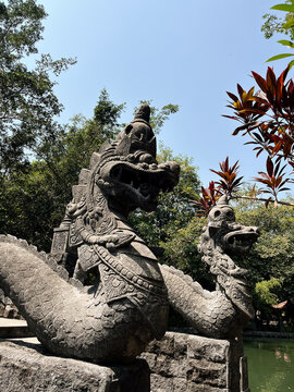 Ancient stone dragon statues guard a tranquil garden pond with lush green trees and vibrant foliage under a clear blue sky.