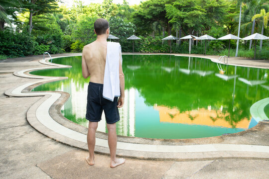 Man seen from behind with towel on shoulder looking at green algae water of abandoned resort pool surrounded by tropical trees and sun umbrellas, disappointed tourist, expectation versus reality