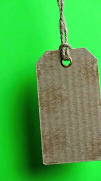 Blank rustic cardboard gift tag swinging gently on a twine string against a bright green screen background for chroma key compositing
