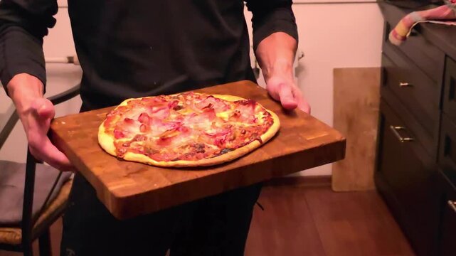 Person presents freshly baked meat pizza on wooden board in home kitchen. Homemade cooking and food preparation. Italian cuisine, savory meal. Close-up of pizza toppings