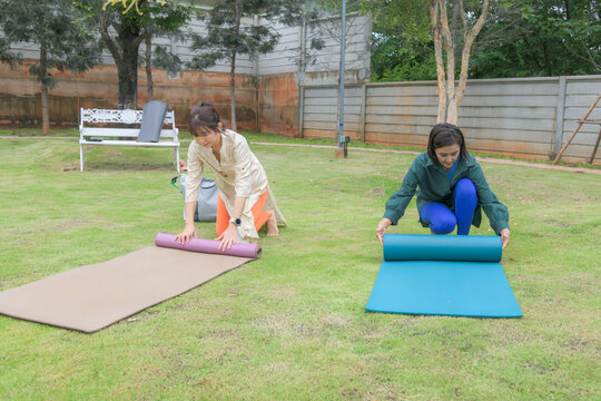 Asian woman in tunic and orange leggings unrolling a beige yoga mat on green grass in a backyard garden while her friend sets up another mat nearby.