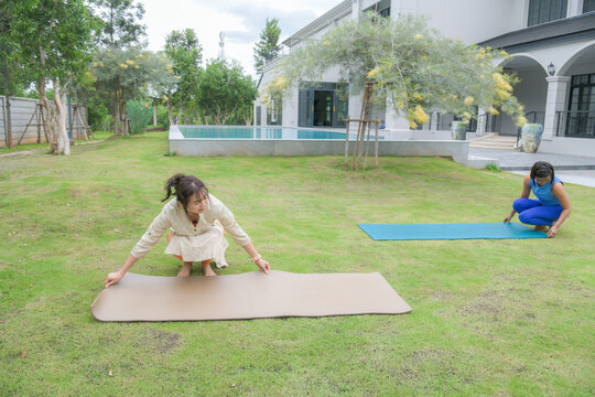 Asian woman in tunic and orange leggings unrolling a beige yoga mat on green grass in a backyard garden while her friend sets up another mat nearby.