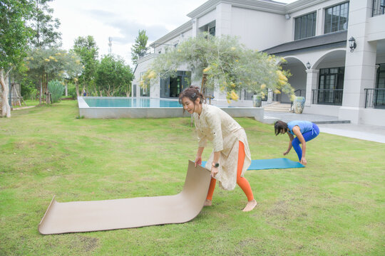 Two Asian women rolling out yoga mats on green lawn outdoors