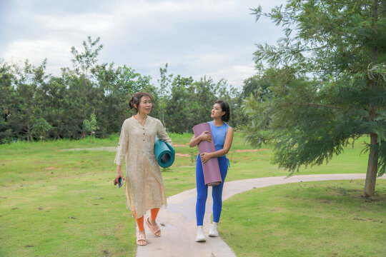Two Asian women walking together along garden pathway with yoga mat