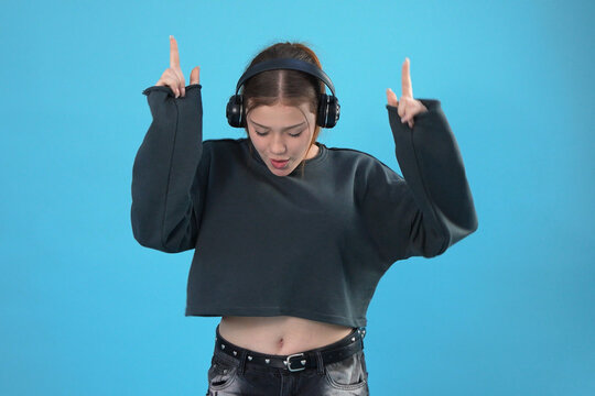 Teen girl listening to music in headphones pointing fingers up against blue background