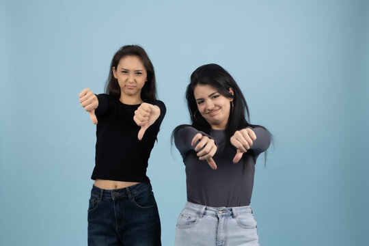 Two Disappointed Young Women Showing Thumbs Down Gestures on Blue Background