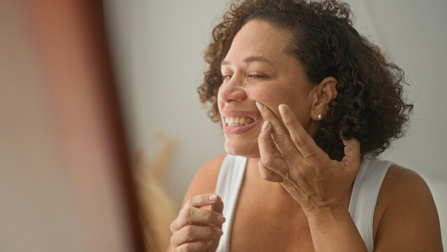Middle aged woman pinches cheek and inspects mouth and teeth in a studio mirror while touching jaw and opening mouth to check alignment; concern selfcare.