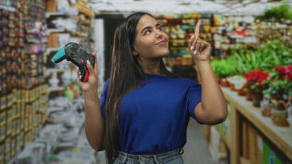 Woman holding gamepad points index finger upward in building aisle lined with plants and shelves, wearing blue shirt and denim jeans  playful confidence. © Krakenimages.com