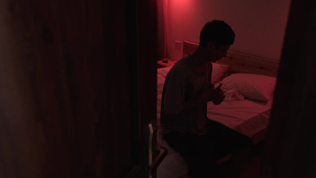 Stressed man sitting alone in a bedroom through a doorway, nervously cracking his fingers and coping with psychological pressure and an uncertain situation in dark low key cinematic light