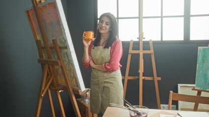 Woman holding orange mug in hand, wearing apron and studying canvas at easel in studio  creative calm. © Krakenimages.com