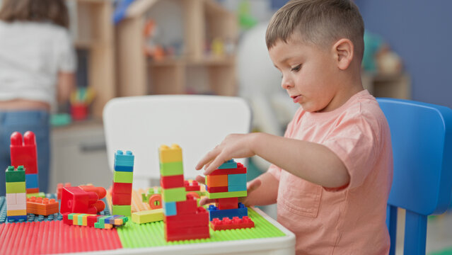 Boy carefully stacking colorful plastic blocks on a play table with hands visible in a building playroom filled with toys and shelves; concentration play.
