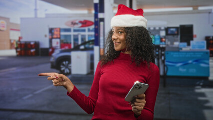 Woman wearing santa hat holds smartphone and points finger while smiling in building entrance  holiday cheerful. © Krakenimages.com