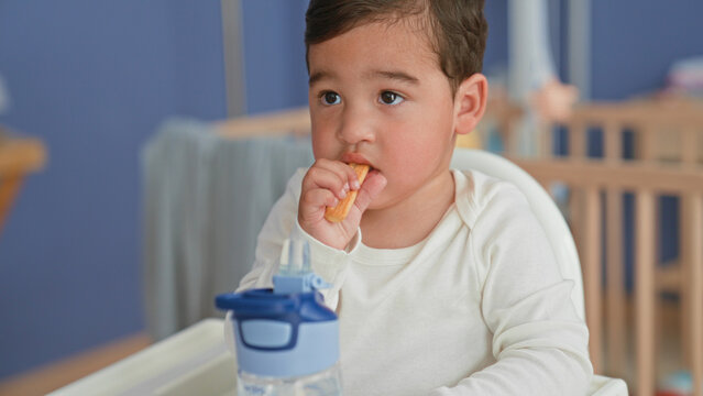 Boy holding cracker to mouth with hand in home nursery highchair beside crib, sippy cup on tray; gentle curiosity.