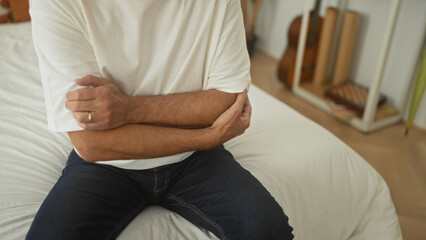 Man clutching left elbow while seated on a bed in a softly lit bedroom inside a residential building  discomfort. © Krakenimages.com