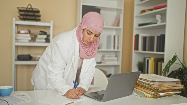 Woman in white coat and pink hijab leaning over laptop writing notes in a cluttered office with stacked files and books in building; professional focus.