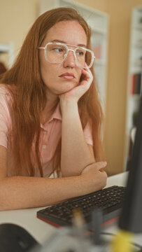 Woman with glasses resting chin on hand at computer keyboard in a home office, freckled face visible and pink polo; boredom reflection.