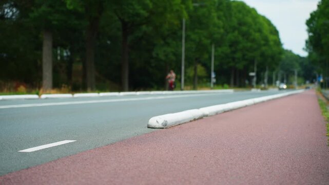 Low-angle view along a red protected cycle path in the Netherlands, separated from the road by a raised white divider with a distant cyclist beneath rows of trees.
