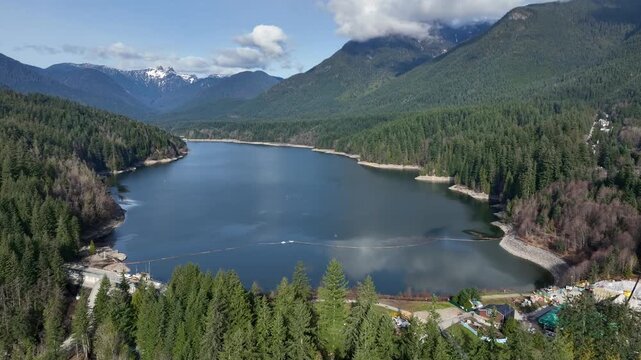 Capilano Lake Dam Basin Framed By Dense Coniferous Forests And Distant Snowcapped Peaks In North Vancouver, British Columbia, Canada. - aerial shot