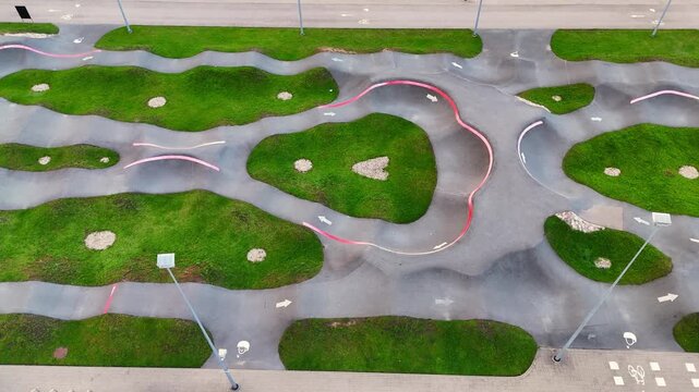 Aerial drone view of a large empty pump track with winding asphalt lanes, banked turns, directional arrows and irregular green islands forming a modern cycling course.