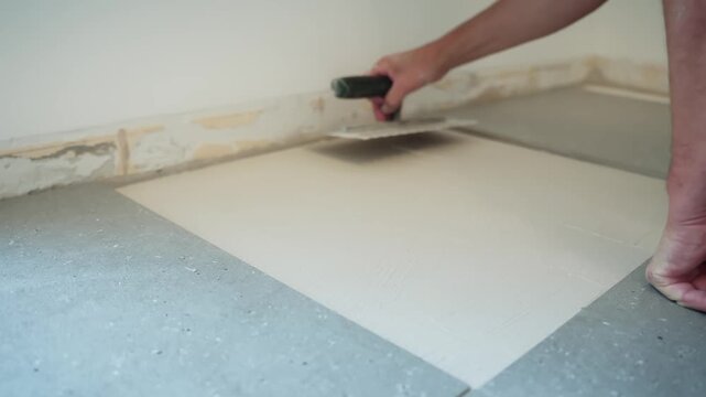 Close-up of a worker smoothing fresh tile adhesive across a floor substrate with a trowel, preparing an even bonding bed before laying the next large-format tile.