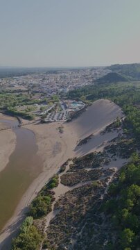 Aerial view of a winding river beside a vast sand dune. Lush greenery borders the sandy slope and calm waters. Town sprawls below, blending into the natural landscape