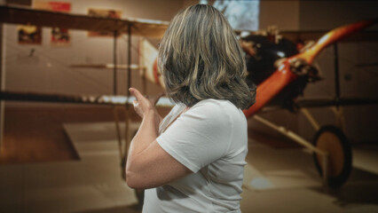 Senior woman in white tshirt finger pointing to vintage biplane display in museum building  nostalgia wonder. © Krakenimages.com